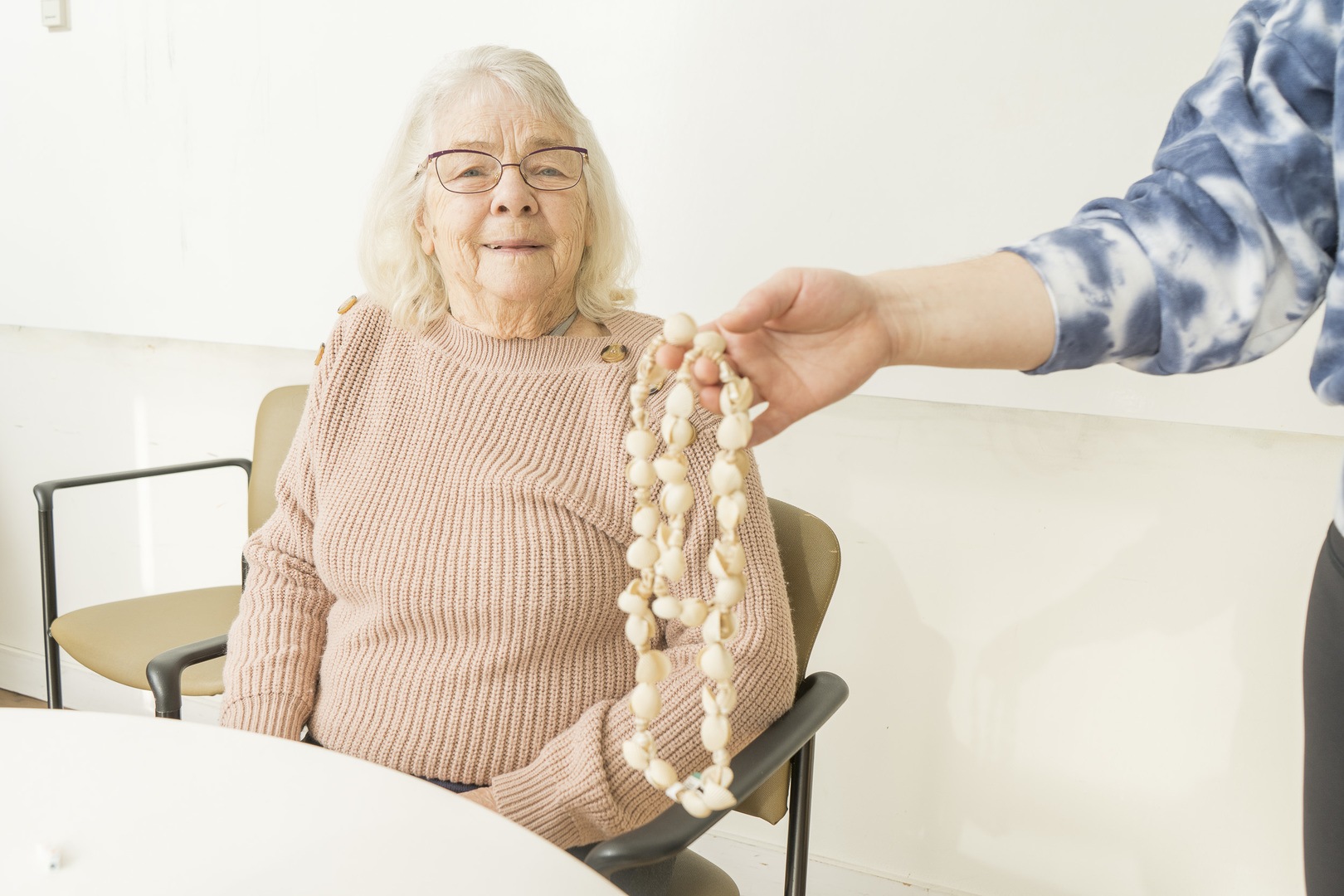 A caregiver hands a senior woman a beaded necklace