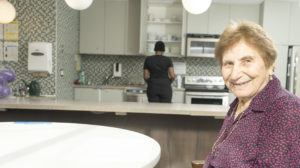 A senior woman sits at a table smiling with her caregiver behind her